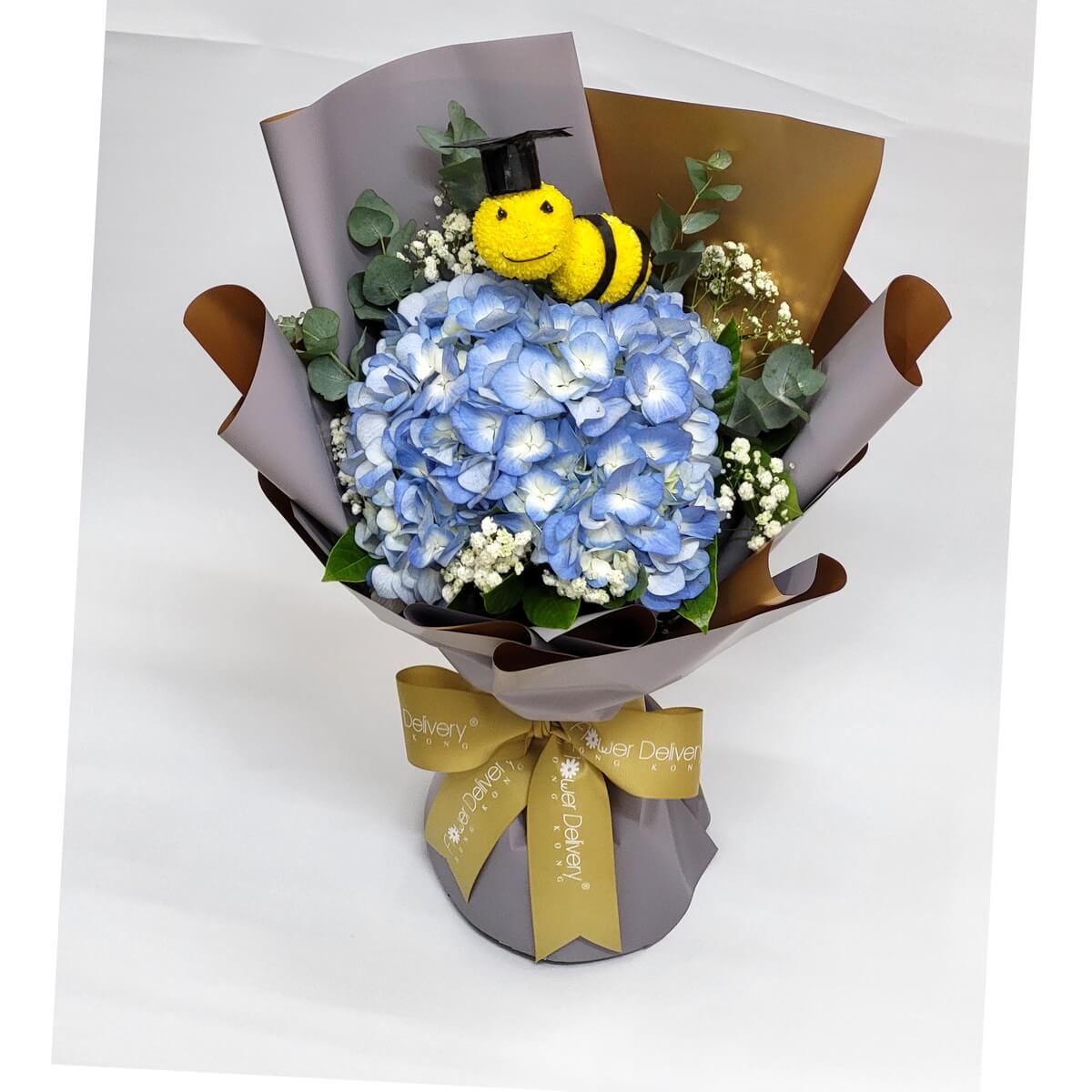 Arrangement of blue hydrangeas, baby’s breath, and eucalyptus with a yellow smiley bee pompom in graduation cap, wrapped in brown-grey paper. 藍繡球花、滿天星、尤加利葉配畢業帽笑臉蜜蜂乒乓菊,灰啡包裝。