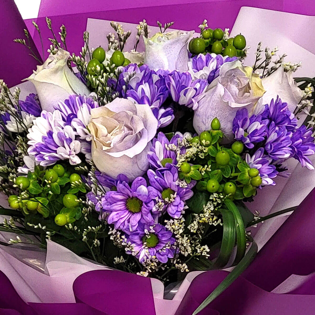 Arrangement of lavender roses, purple mums, green berries, and white fillers 薰衣草色玫瑰與紫色小菊、綠色鼠尾草果交織,點綴白色星點配花,搭配紫白包裝紙,整體柔美而富層次感。