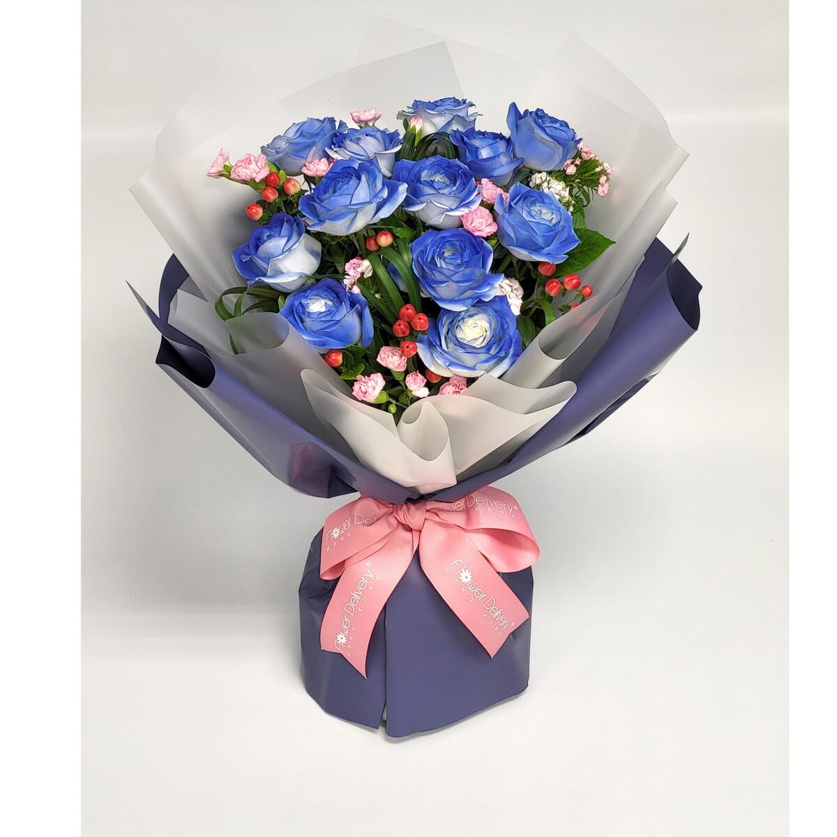 Arrangement of blue roses, pink mini carnations, red hypericum berries, and folded greenery, wrapped in white and navy paper with a pink ribbon. 藍玫瑰、粉紅迷你康乃馨、紅豆花及綠葉,白色與深藍包裝,粉紅絲帶裝飾。