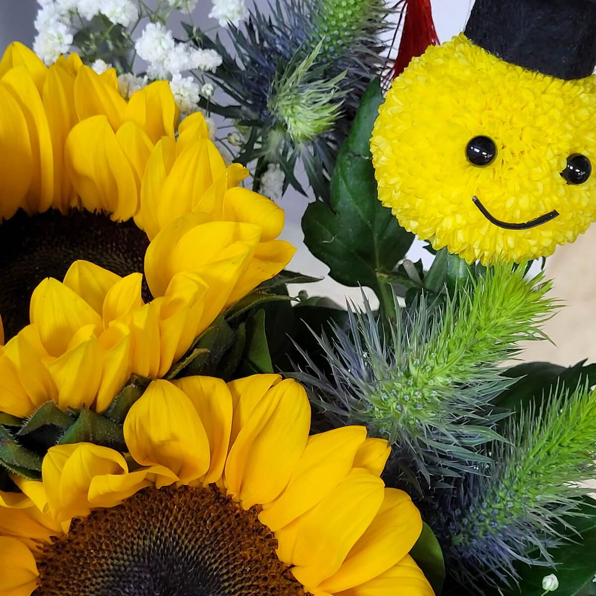 Detail view of vibrant sunflowers, blue eryngium, white statice, and a graduation-themed yellow button chrysanthemum smiley face 向日葵、藍松球、白紗與畢業主題黃色乒乓菊笑臉造型花藝細節特寫
