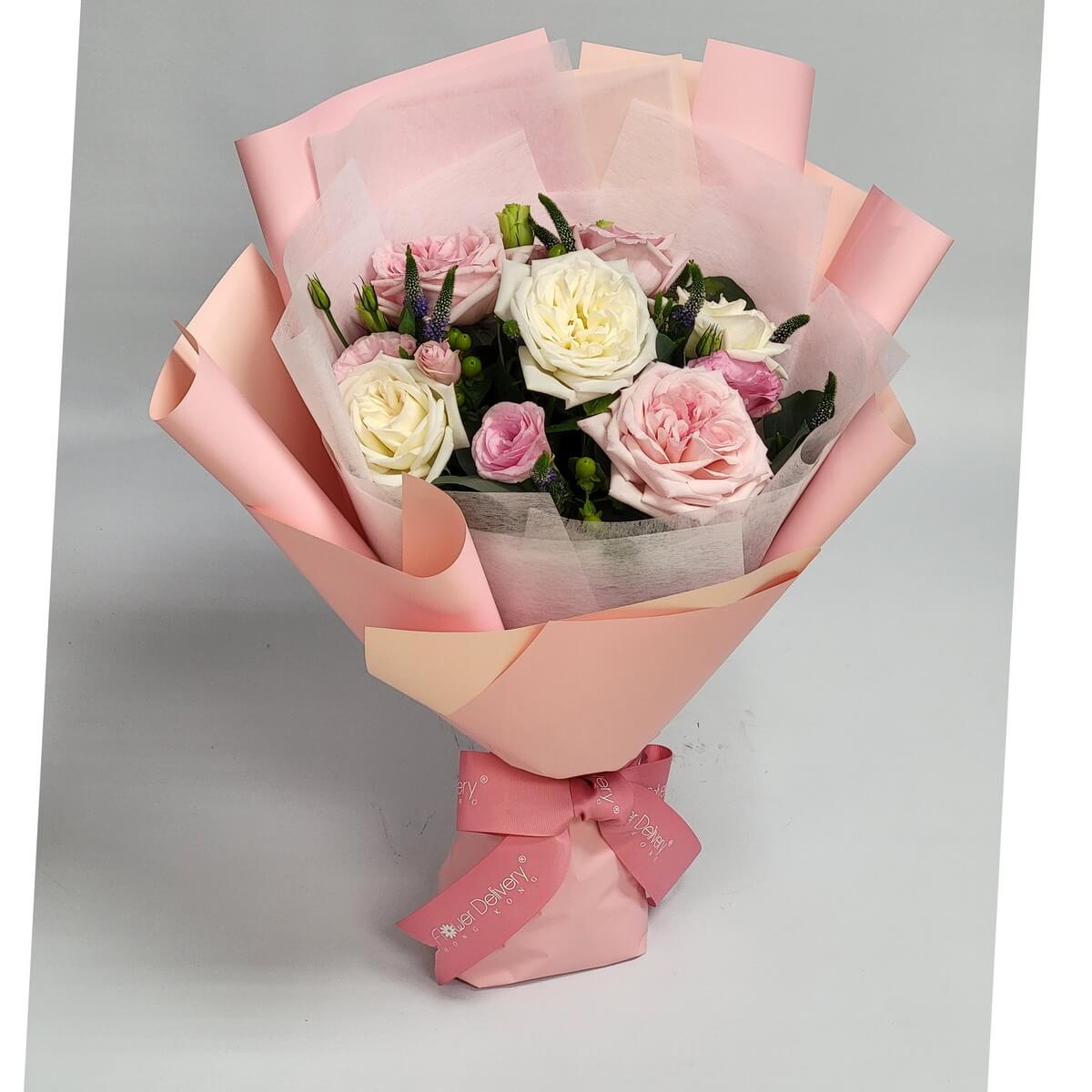 A pastel bouquet of blush pink and creamy white roses, with soft greenery, wrapped in pale pink paper and tied with a pink ribbon. 粉紅與奶白玫瑰搭配嫩綠葉材,粉色包裝展現甜美與柔情。