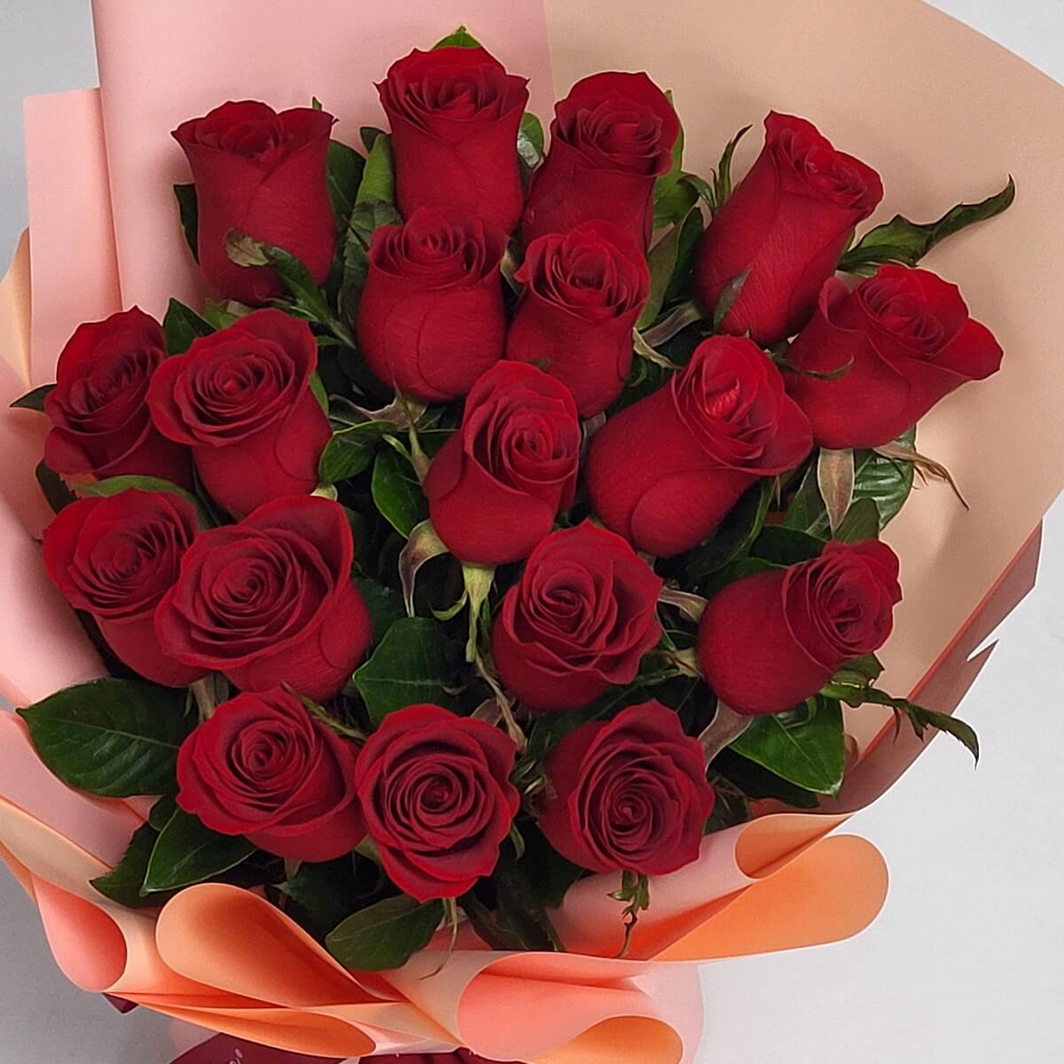Full bouquet featuring red roses in tight formation, accented with lush green leaves and wrapped in curled pink and peach paper for a clean, modern look - 紅玫瑰密集排列,配綠葉,粉紅及蜜桃色包裝花束,造型俐落時尚