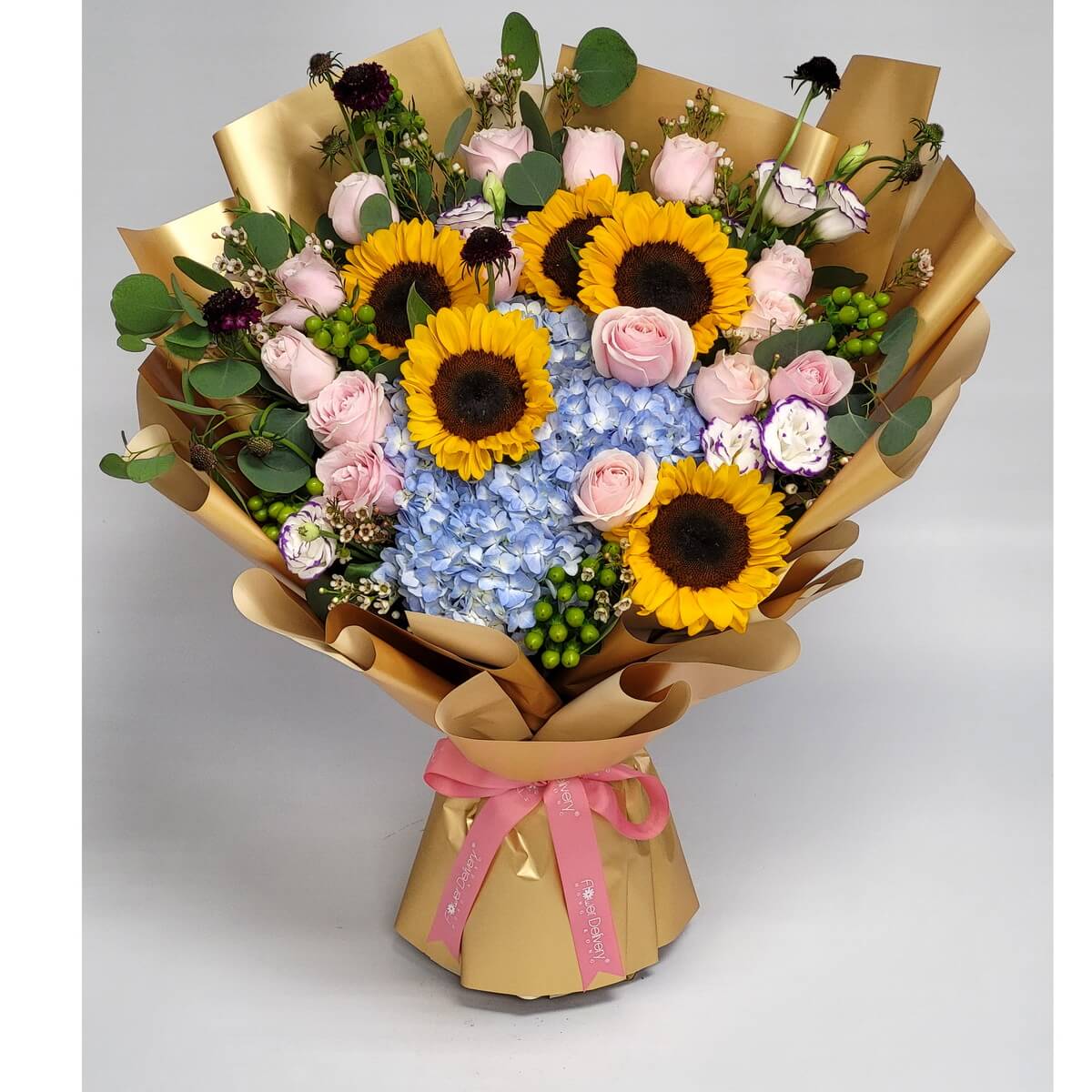 Vivid bouquet of sunflowers, pink roses, and blue hydrangeas wrapped in gold with a pink ribbon. 明亮向日葵搭配粉紅玫瑰與藍繡球,金色包裝與粉紅絲帶散發溫馨祝福。