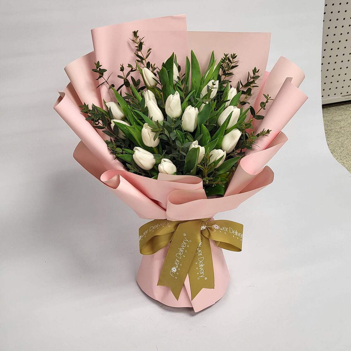 A minimalist bouquet of pure white tulips with lush greenery, wrapped in soft blush pink paper and tied with a gold ribbon. 清新脫俗的氣質,展現簡約優雅之美。