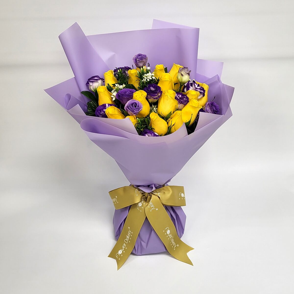 A bold bouquet of vibrant yellow roses and purple eustomas, accented with hints of white blooms and greenery. Wrapped in soft lavender paper with a gold ribbon. 黃玫瑰與紫洋桔梗交織成搶眼對比,展現自信與優雅。