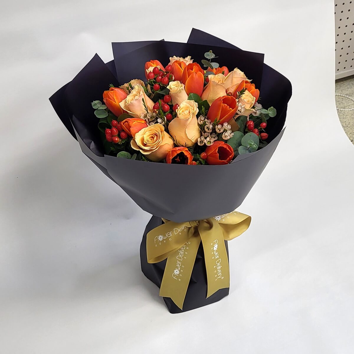 A rich bouquet of orange tulips, champagne roses, red hypericum berries, and greenery, wrapped in elegant matte black paper with a gold ribbon. 橙色鬱金香、香檳玫瑰與紅色冬青果交織,搭配墨黑包裝與金緞帶,展現秋意與高貴風格。