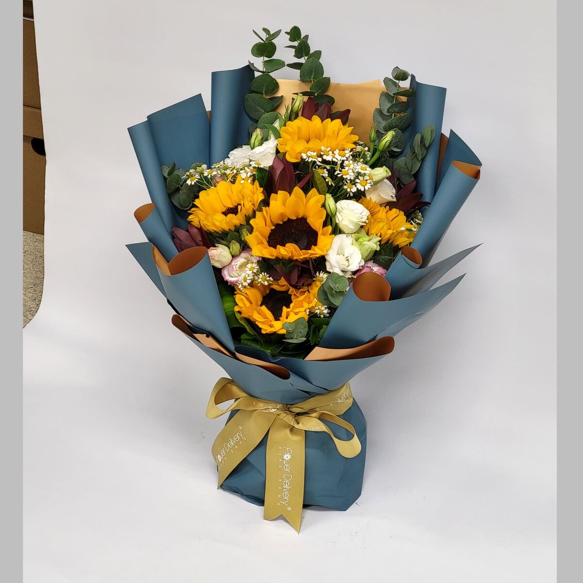 A joyful bouquet featuring bright sunflowers, white lisianthus, chamomile, and eucalyptus, wrapped in deep teal and gold paper. 向日葵搭配洋桔梗、洋甘菊與尤加利葉,深藍與金色包裝,洋溢陽光氣息。