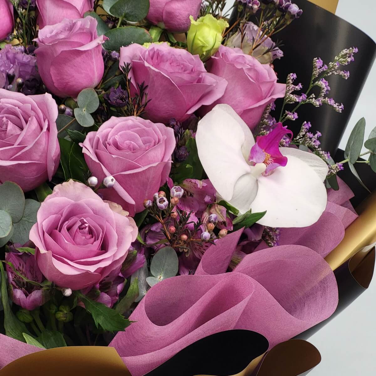 Detailed view of mauve roses accented by a white Phalaenopsis orchid, pink wax flowers, eucalyptus leaves, and delicate lavender blooms in pink mesh wrap - 粉紫玫瑰配白蝴蝶蘭、粉蠟花、尤加利葉及薰衣草花,粉色網布包裝特寫