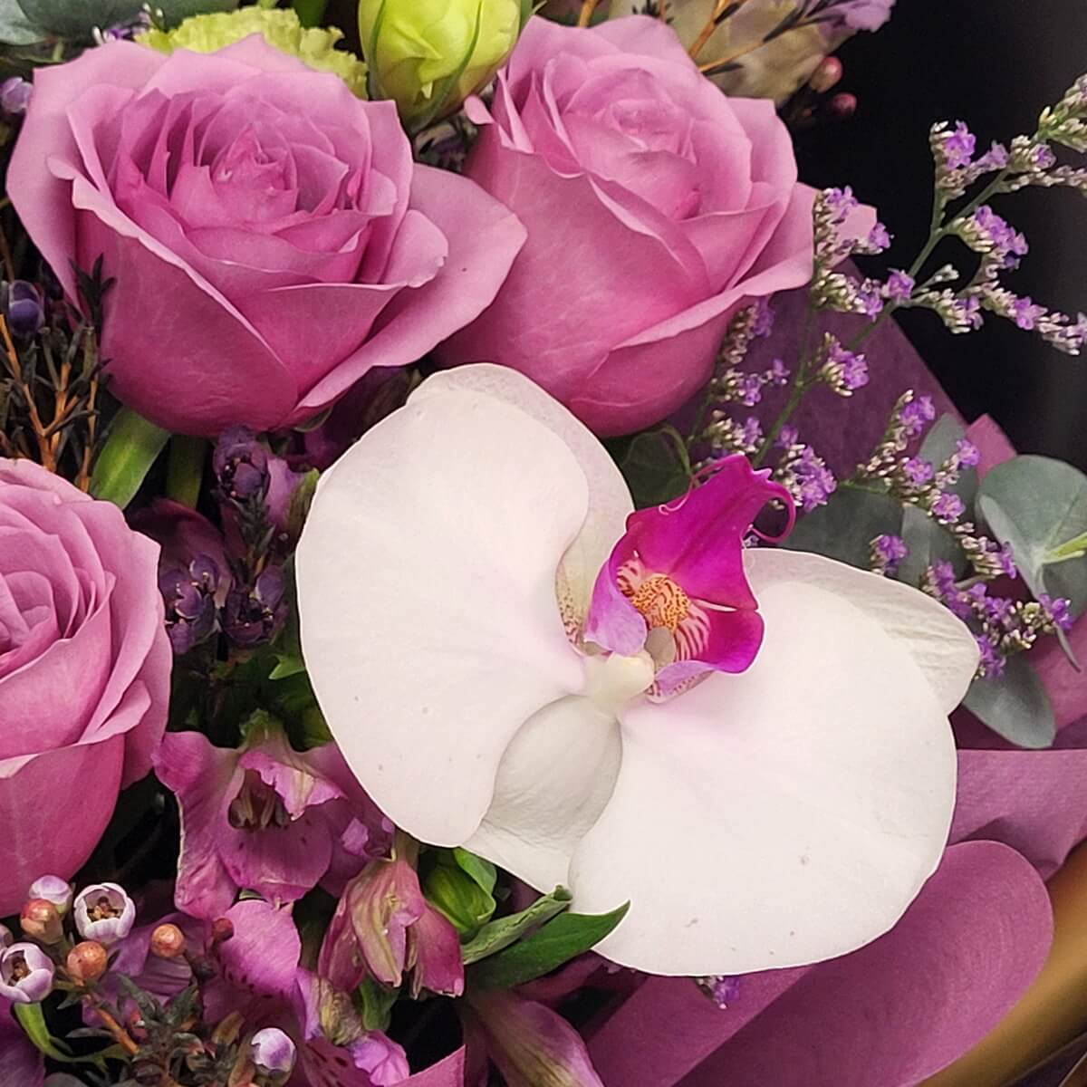 Detail view of a white Phalaenopsis orchid with purple center, surrounded by mauve roses, pink wax flowers, statice, and eucalyptus foliage - 白蝴蝶蘭(紫紅唇瓣)配粉紫玫瑰、粉蠟花、紫丁香及尤加利葉特寫