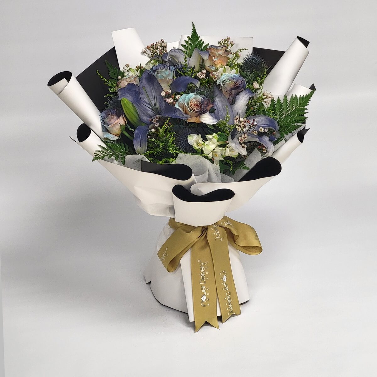Bouquet of muted rainbow roses and deep indigo calla lilies, paired with greenery and white wax flowers, wrapped in modern black-and-white paper with a champagne gold ribbon. 暗彩玫瑰與靛紫馬蹄蓮搭配白色蠟花與綠葉,黑白包裝展現時尚魅力。