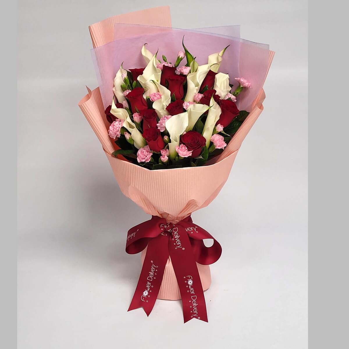 A graceful bouquet of white calla lilies, red roses, and pink mini carnations, arranged in a soft champagne pink ribbed wrapper with translucent pink accents. Tied with a bold red ribbon. 高貴的馬蹄蓮與紅玫瑰交織,展現莊重與愛戀。