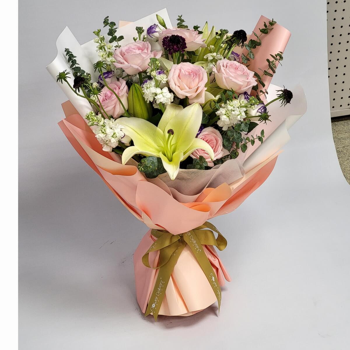 Elegant bouquet featuring soft pink roses, white lilies, purple scabiosas, and seasonal blooms, wrapped in layers of champagne and blush paper with a moss green ribbon. 粉玫瑰與百合交織紫花,襯托夢幻香檳包裝,溫柔高貴。