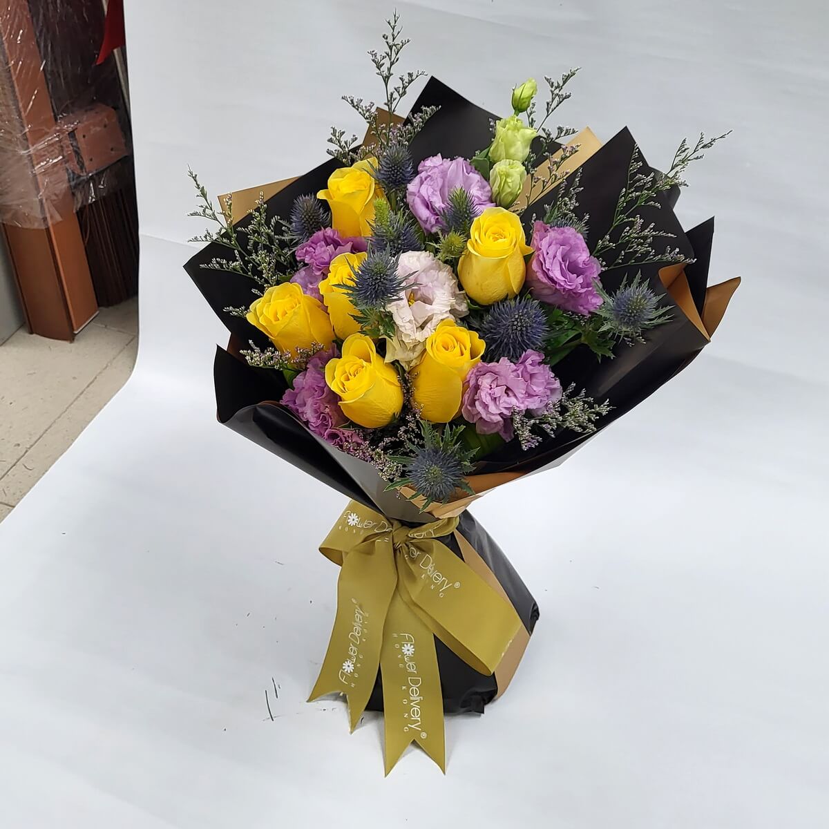 A vibrant bouquet of yellow roses, purple eustomas, and spiky blue thistles, accented with pale greenery. Wrapped in sleek black and gold paper with a gold ribbon. 鮮黃玫瑰、紫色桔梗與藍蓟花相襯,黑金包裝展現時尚高貴氣息。