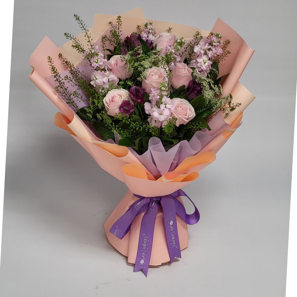 A delicate bouquet of pale pink roses, light mauve stock, and purple alstroemerias, surrounded by soft greenery. Wrapped in peach and lavender-toned paper with a purple ribbon. 淡粉玫瑰、淺紫紫羅蘭、紫色百合草與綠葉交織,包裹於蜜桃與薰衣草色包裝紙中,優雅迷人。