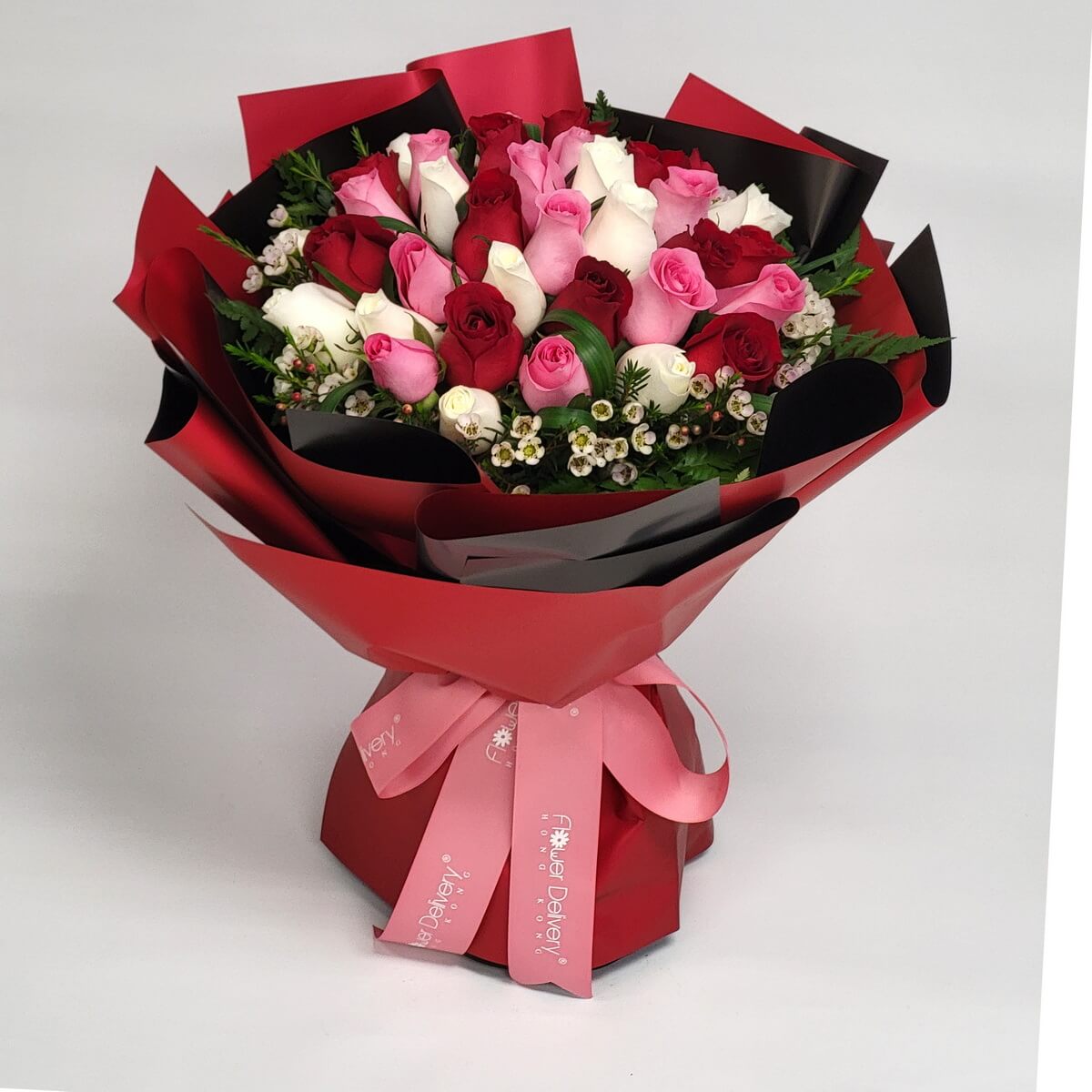 A romantic bouquet of red, pink, and white roses arranged in a circular pattern, accented with waxflowers and wrapped in bold red and black paper with a pink ribbon. 紅、粉、白玫瑰交錯排列,搭配蠟花,紅黑包裝與粉紅絲帶增添濃情氣息。