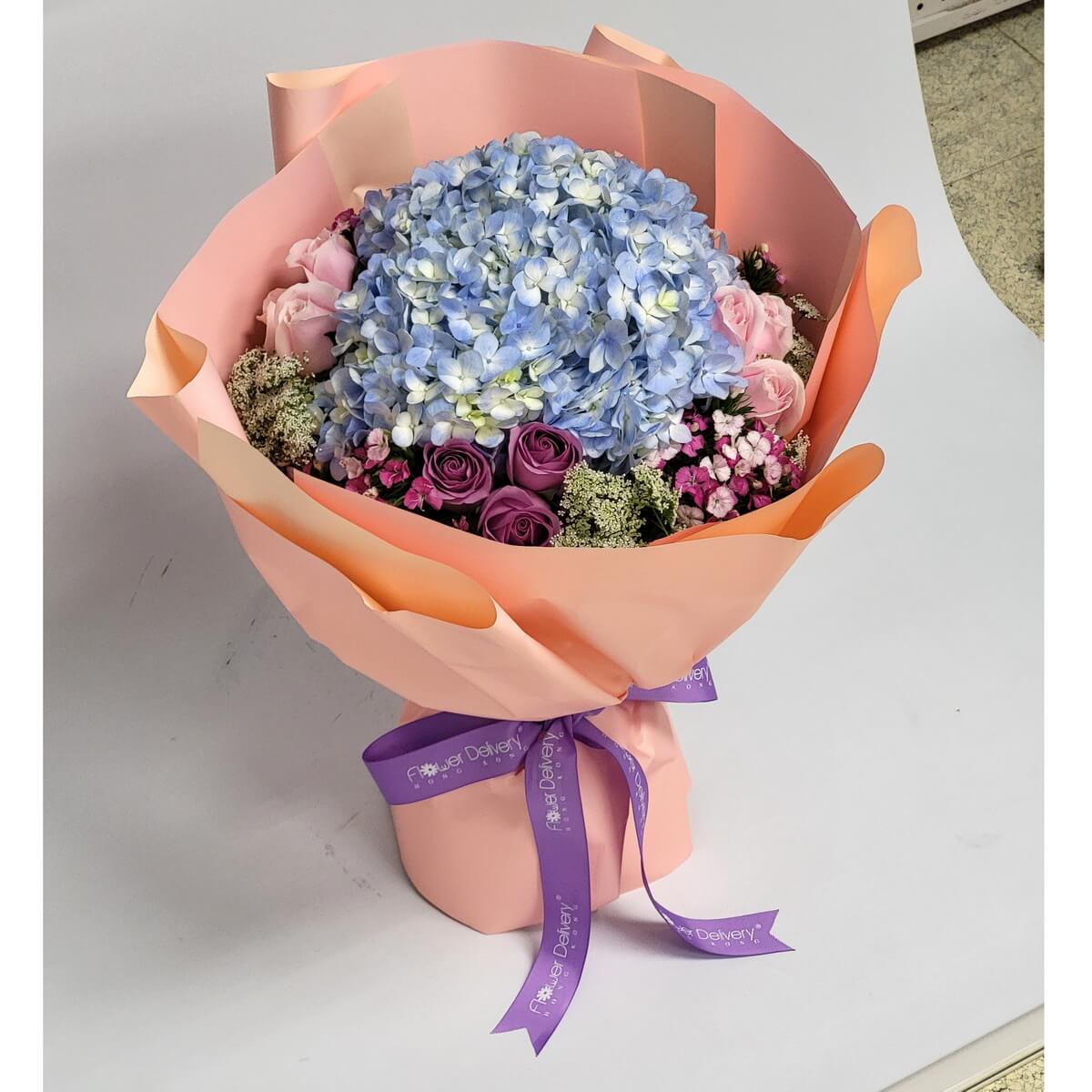 A graceful bouquet featuring a large blue hydrangea surrounded by lavender roses, pink roses, and delicate filler flowers, wrapped in peach paper with a purple ribbon. 藍繡球搭配薰衣草玫瑰與粉玫瑰,粉橘包裝與紫絲帶增添優雅氣息。