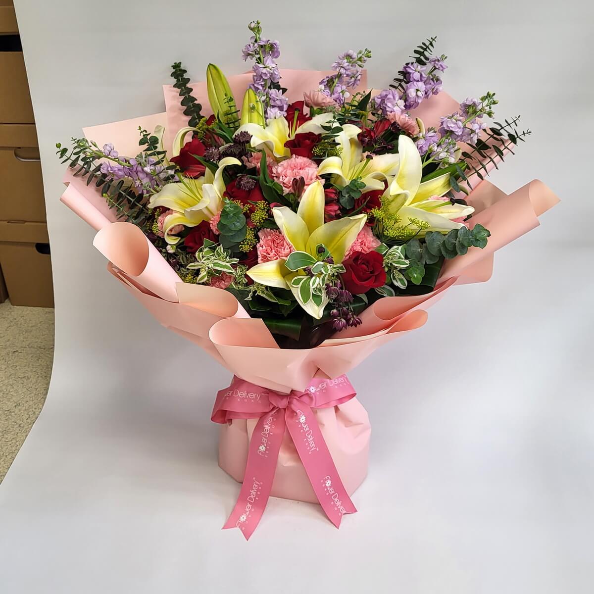 A grand bouquet of champagne lilies, red roses, pink carnations, and purple stock, wrapped in blush pink paper with a matching ribbon. Elegant and festive. 粉紅包裝襯托香檳百合、紅玫瑰、粉康乃馨與紫羅蘭,典雅華麗,滿載祝福。