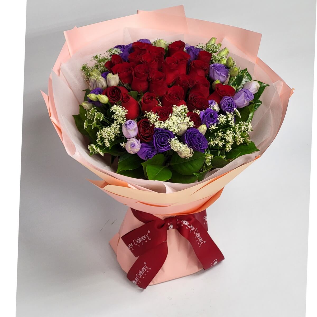 A dramatic bouquet of deep red roses, purple lisianthus, and delicate white filler flowers, wrapped in peach-toned paper with a crimson ribbon. 濃紅玫瑰與紫桔梗交織,佐以白色碎花,桃橘包裝紙與酒紅緞帶襯托出濃烈情感。
