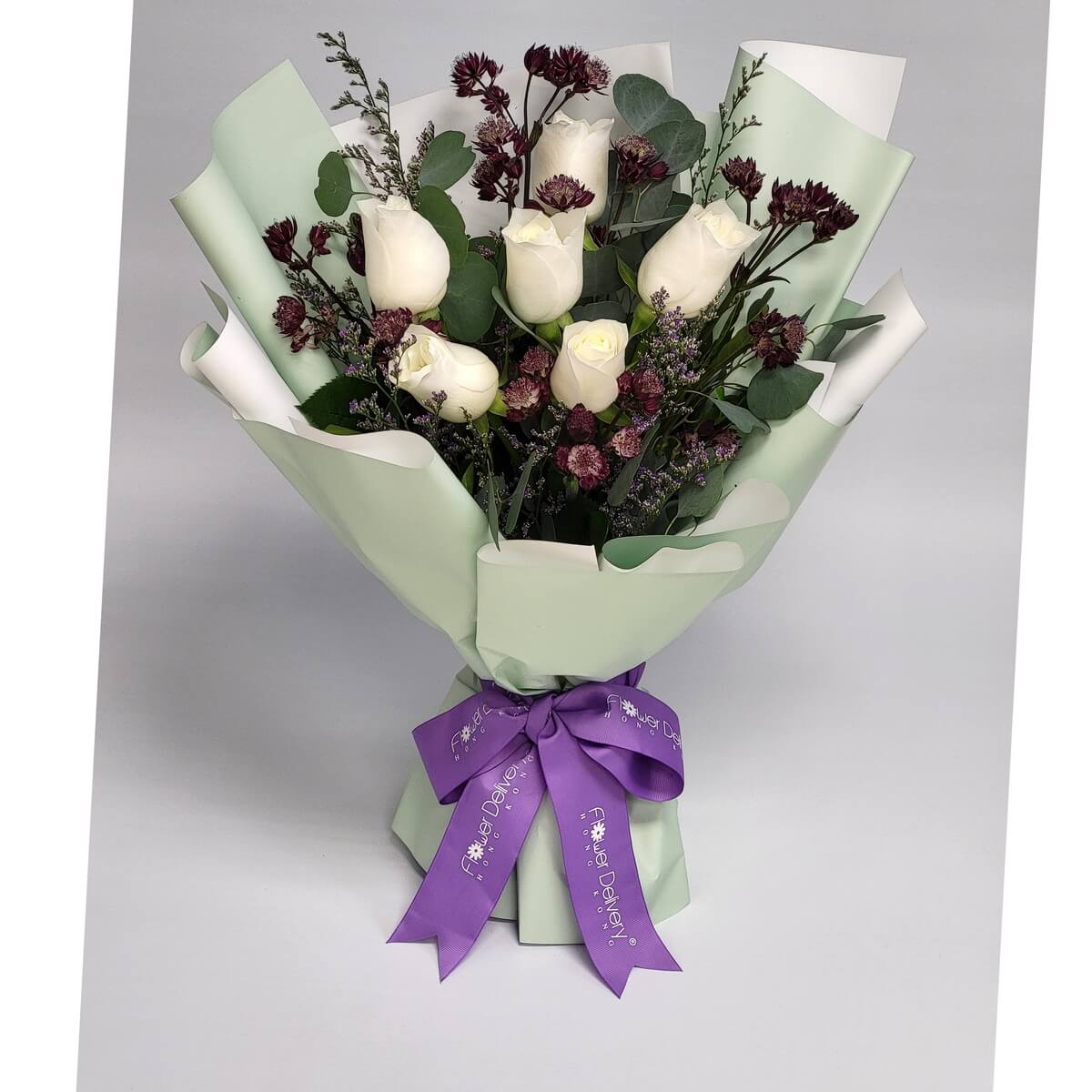 A graceful bouquet of white roses, deep burgundy button flowers, eucalyptus, and lilac filler, wrapped in pale green and white paper with a purple ribbon. 白玫瑰搭配酒紅小花與尤加利,襯以淡綠白包裝與紫色緞帶,展現優雅與靜謐。