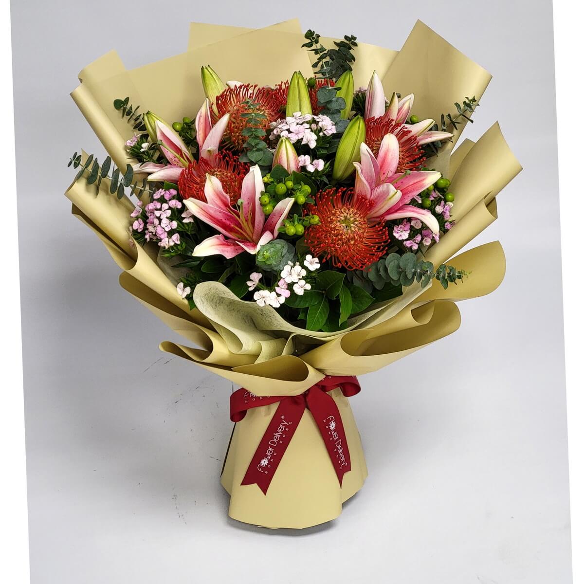 A bold bouquet of pink lilies, red pincushion proteas, pink kalanchoes, green hypericum berries, and eucalyptus, wrapped in champagne gold paper with a red ribbon. 鮮豔粉紅百合與針墊花,綠色尤加利與紅色包裝點綴,充滿熱情活力。