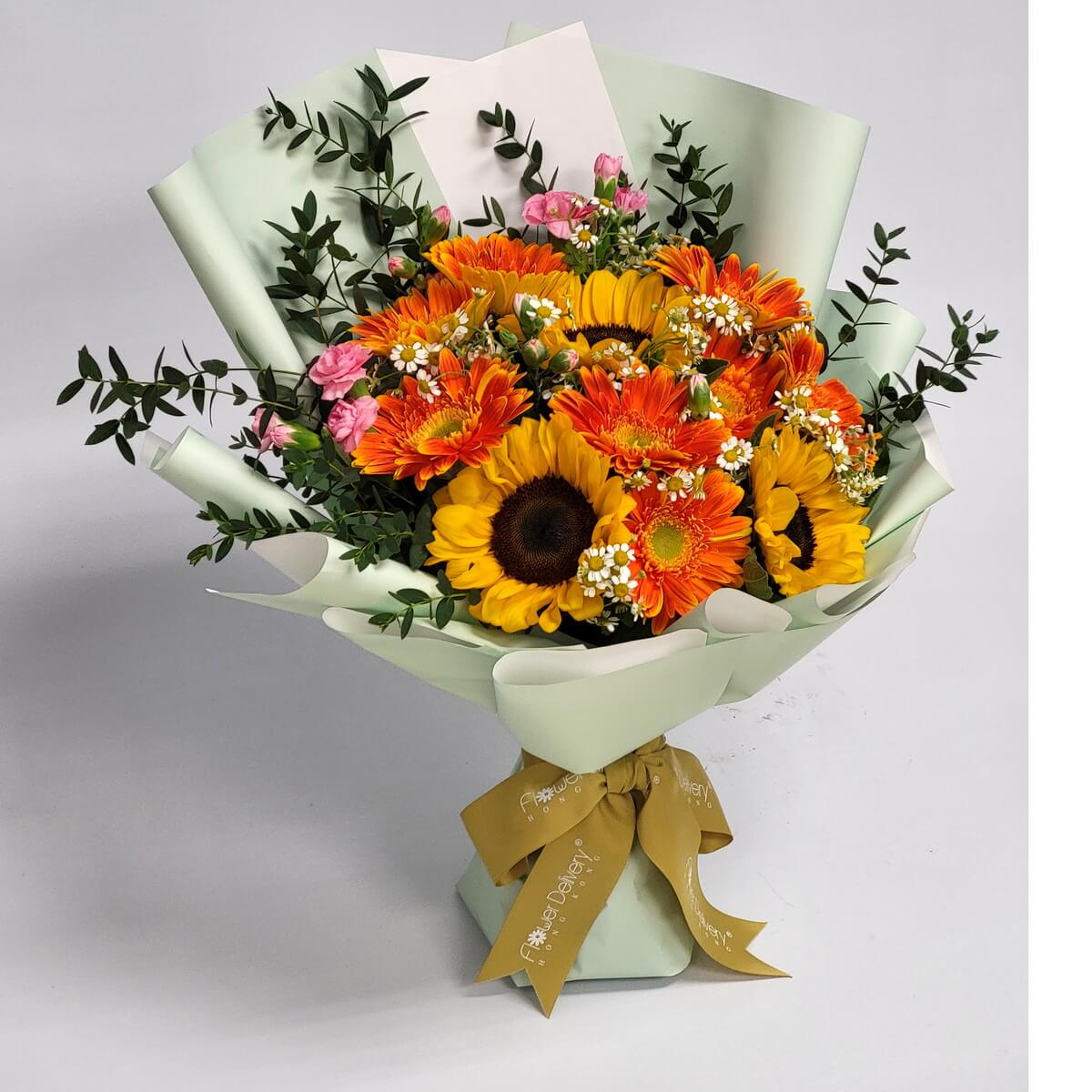 A vibrant bouquet of sunflowers, orange gerberas, pink mini carnations, and baby's breath with lush greenery, wrapped in mint green paper and a mustard yellow ribbon. 向日葵與橘色太陽菊搭配粉紅康乃馨,清新綠色包裝綻放陽光氣息。