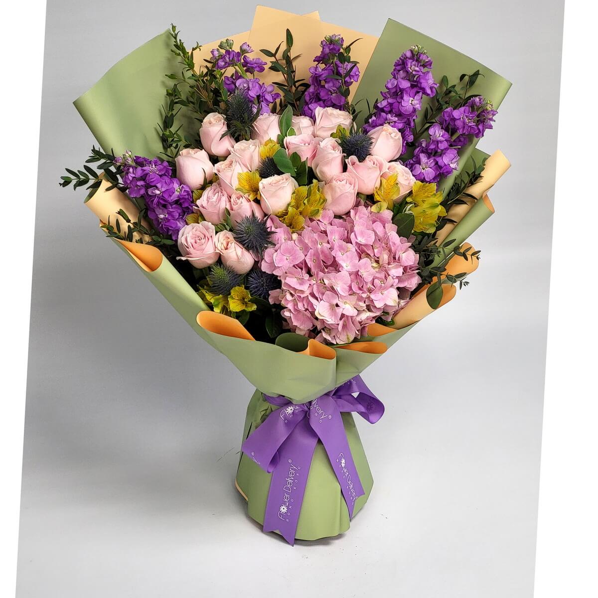 A lavish bouquet featuring pale pink roses, purple stock, pink hydrangea, yellow alstroemeria, and blue eryngium, wrapped in sage green and champagne paper with a purple ribbon. 粉玫瑰、紫花與粉繡球交織成華麗花束,綠金包裝添上高雅氣息。