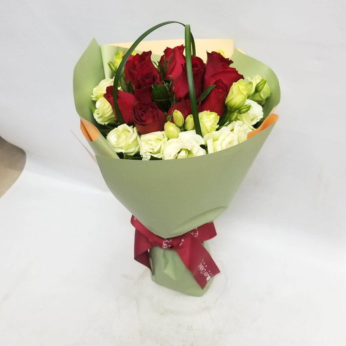 A bouquet of classic red roses surrounded by creamy white lisianthus, wrapped in sage green paper and tied with a deep red ribbon. 清新綠包裝內紅玫瑰與奶白洋桔梗交織,展現浪漫與沉穩的對比。