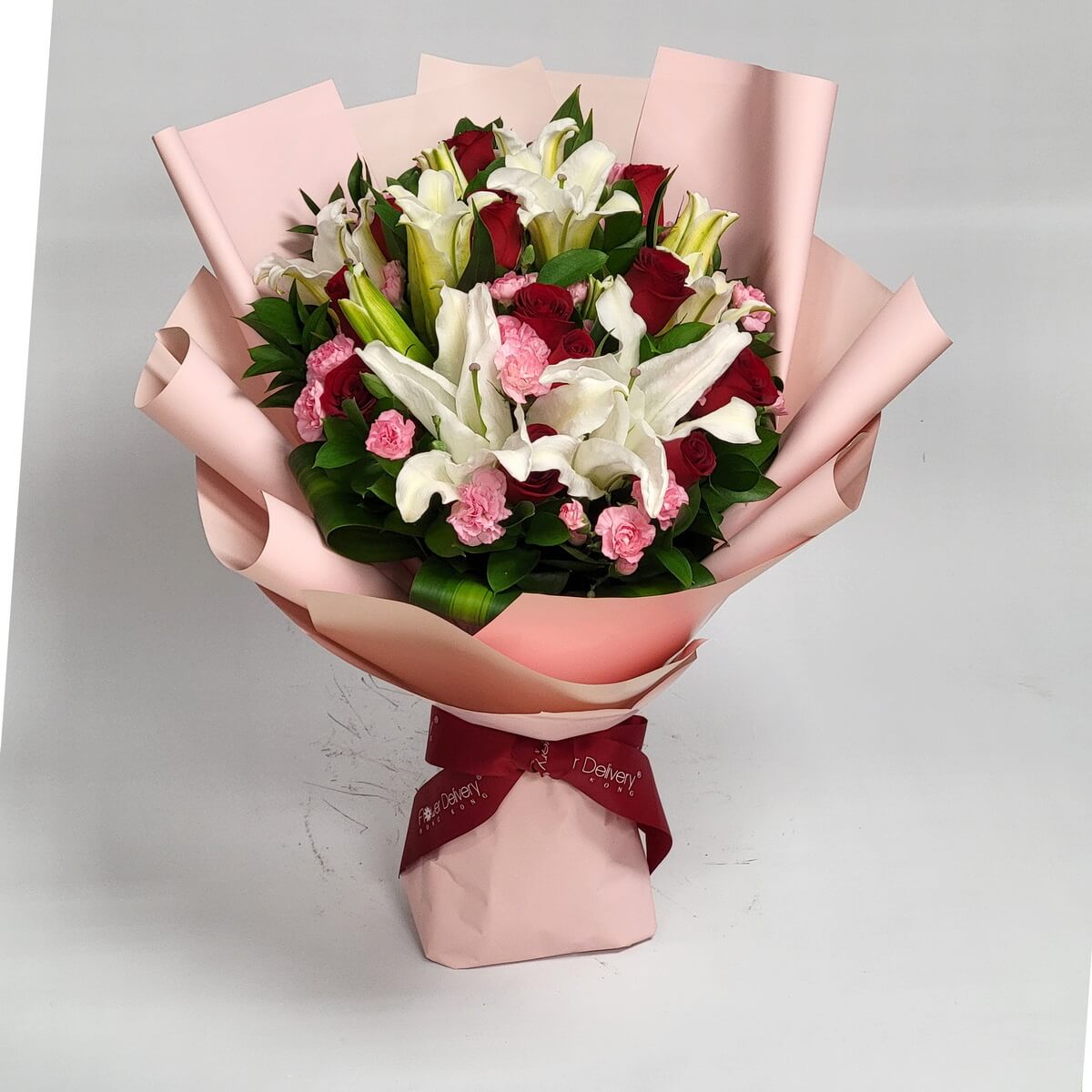 Bouquet of white lilies, red roses, and pink carnations, wrapped in soft pink and champagne paper with a deep red ribbon. Graceful and romantic. 百合、紅玫瑰與粉康乃馨混合,粉香檳色包裝,酒紅絲帶點綴,優雅浪漫。