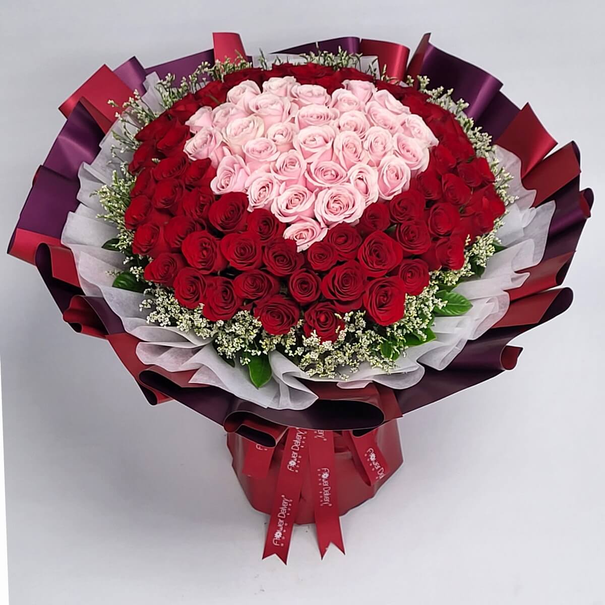 Heart-shaped arrangement of soft pink roses framed by deep red roses, wrapped in layered burgundy and white paper with red ribbons. 心形粉紅玫瑰搭配紅玫瑰,包裹紫紅與白色層次包裝紙,浪漫真摯。