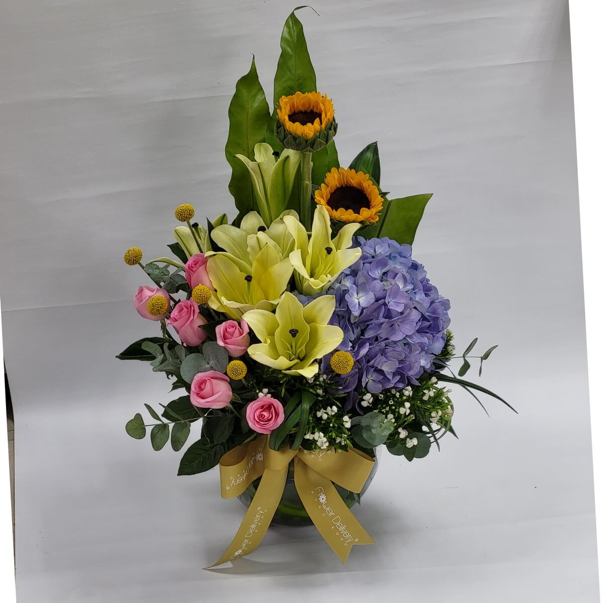 Tall floral design with sunflowers, yellow lilies, purple hydrangea, pink roses, craspedia, and baby's breath in a round glass vase with champagne ribbon 向日葵、黃百合、紫繡球、粉玫瑰、金球花與滿天星,搭配綠葉,層次分明,氣氛明亮愉悅。