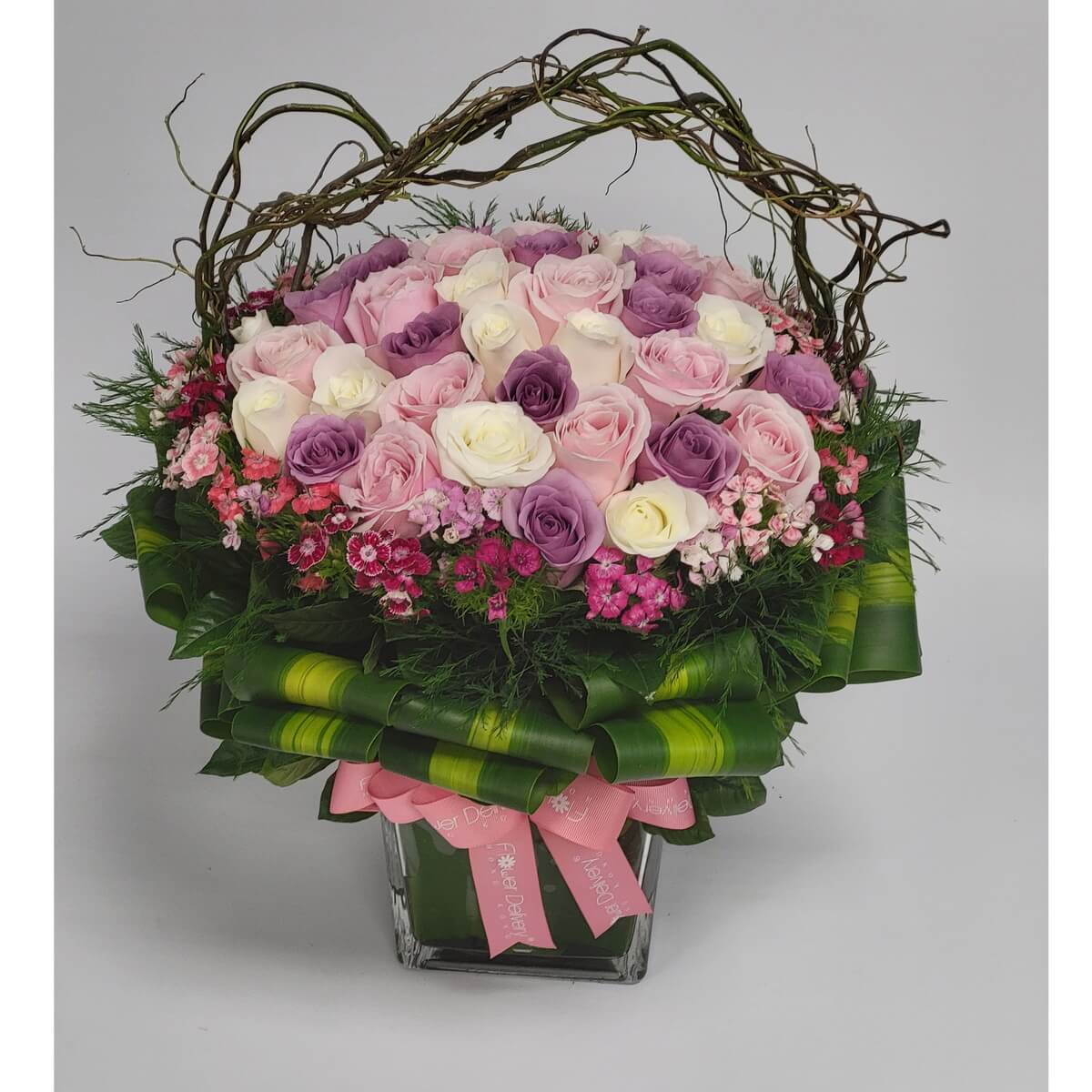 Lavender, blush, and white roses in lush design 淡紫、粉紅與白玫瑰,搭配迷你花朵與綠葉層層圍繞,藤蔓拱形點綴,方形花瓶綁以粉紅緞帶,展現典雅與浪漫氣息。