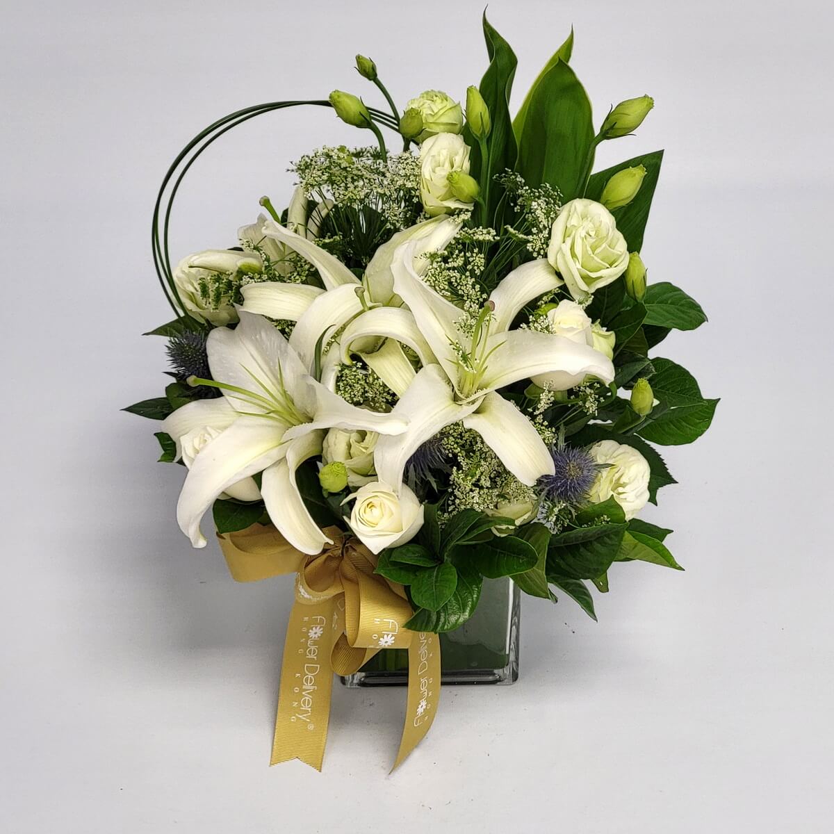 White lilies, roses, lisianthus and soft greenery in a square vase with gold ribbon 純白百合、玫瑰與洋桔梗交織出清新優雅,搭配金色緞帶與綠葉點綴,呈現出高貴靜謐的氛圍。