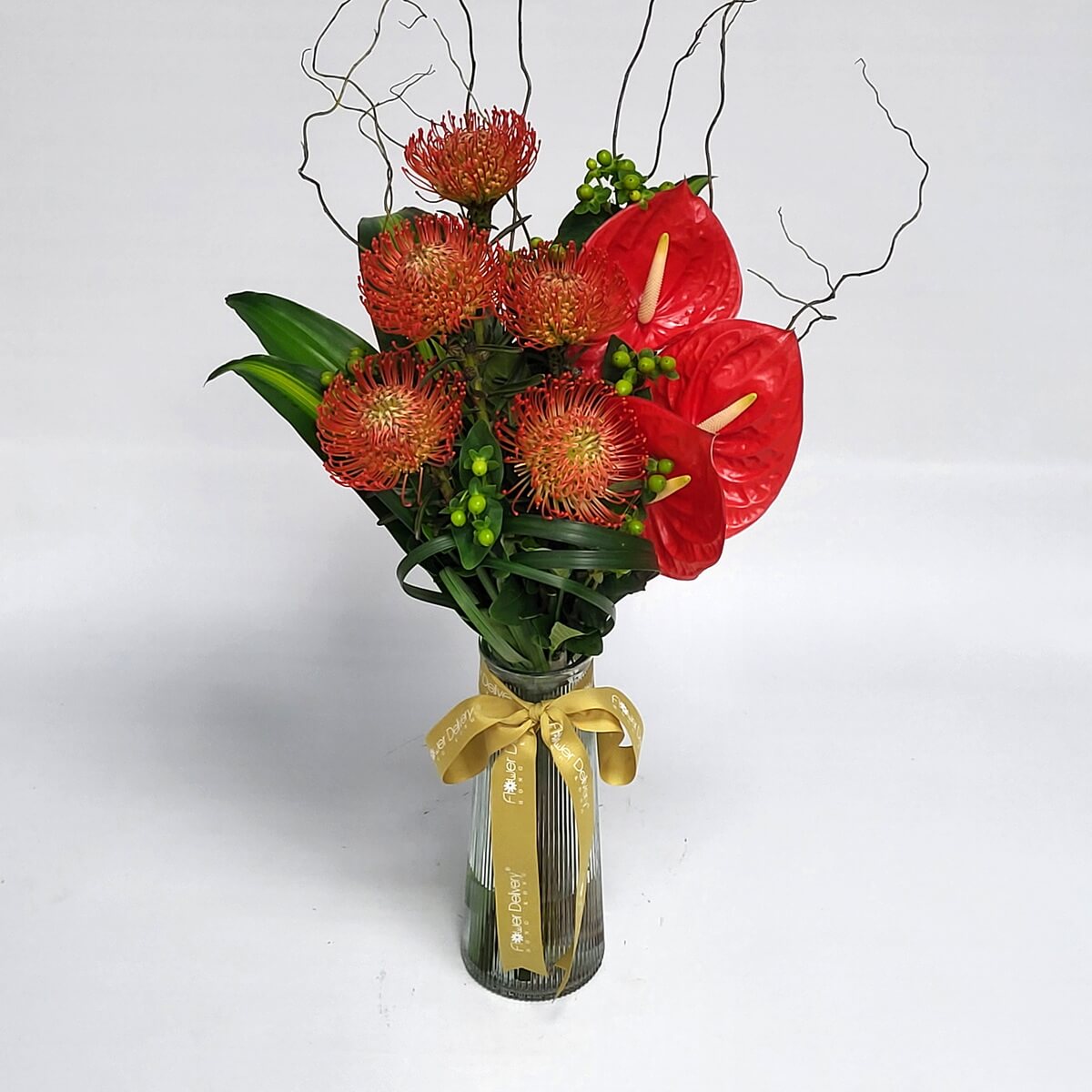 Striking red pincushion proteas and anthuriums with curly branches in a tall vase 紅色針墊花配火鶴花與捲曲枝條,設計前衛獨特,置於條紋玻璃瓶中,展現熱帶風情與現代藝術感。