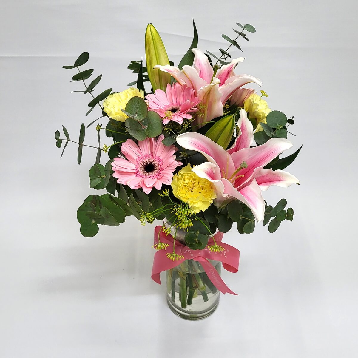 Fresh bouquet of pink and yellow blooms in vase 粉百合、非洲菊、黃康乃馨與圓葉尤加利葉搭配,插於透明玻璃瓶中,繫上粉紅蝴蝶結,展現柔和溫馨氛圍。