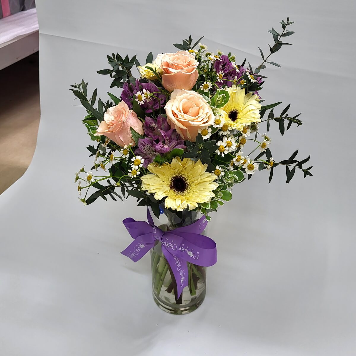 Bright mixed arrangement in vase 玻璃瓶綁紫色絲帶,內含香檳玫瑰、黃太陽菊、紫色紅掌與洋甘菊,綠葉襯托,整體風格清新自然,充滿春日氣息。