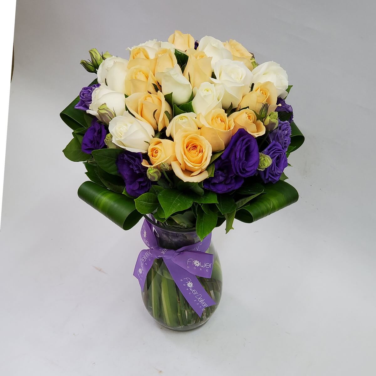 Arrangement of yellow and white roses with deep purple lisianthus in a clear glass vase, accented with foliage – 黃白玫瑰與深紫洋桔梗花束,配綠葉,置於透明玻璃花瓶中
