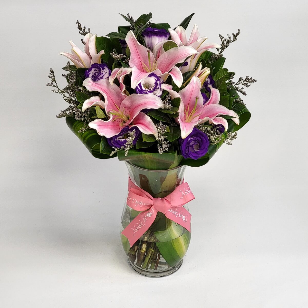 Pink lily and purple eustoma bouquet 粉色百合與紫色桔梗交織成花束,配以熱帶葉材與灰色襯花,裝於曲線玻璃瓶中,粉紅蝴蝶結點綴,展現高雅大方風格。