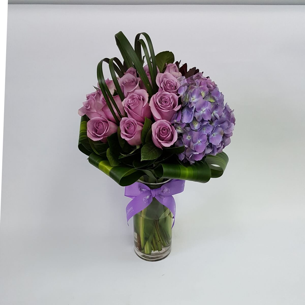 Elegant arrangement of purple roses and hydrangeas, accented with lush greenery in a glass vase 紫玫瑰與紫繡球花搭配繁茂綠葉,玻璃瓶承載,展現高雅氣質。