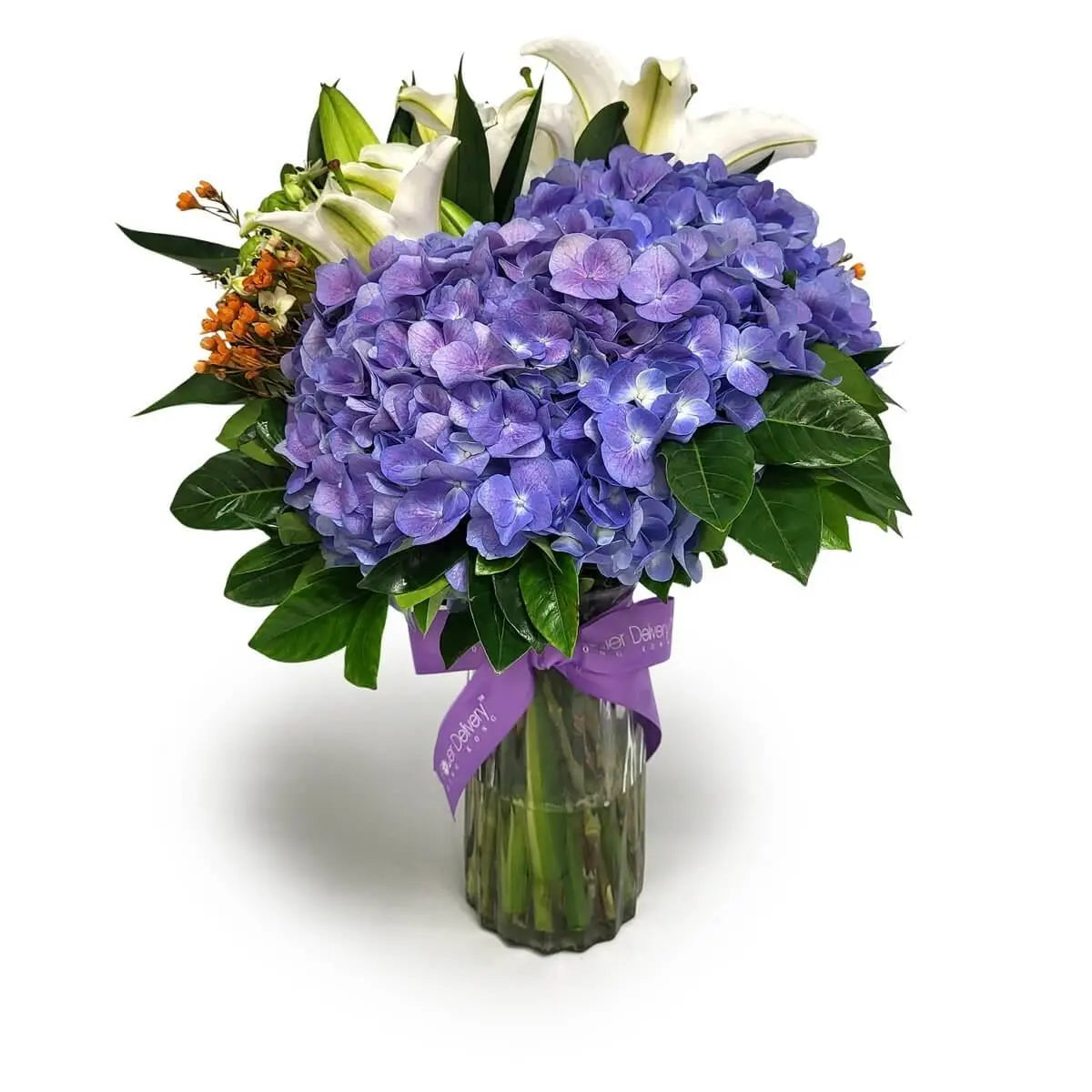 Glass vase arrangement featuring purple hydrangeas, white lilies, orange filler flowers, and dark green foliage, tied with a lavender ribbon. 紫繡球、白百合、橙色配花與深綠葉插於玻璃瓶,綁以紫色絲帶。