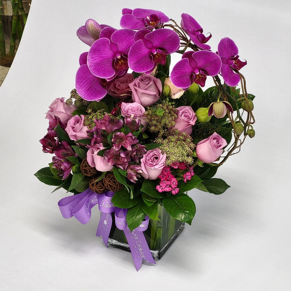 Original image of full floral arrangement with bright fuchsia orchids, lavender roses, green Queen Anne’s lace, pink alstroemeria, spray roses, and decorative foliage in a glass cube vase. 洋蘭、紫玫瑰、蕾絲花、粉百合、小玫瑰與綠葉方形玻璃瓶插花。
