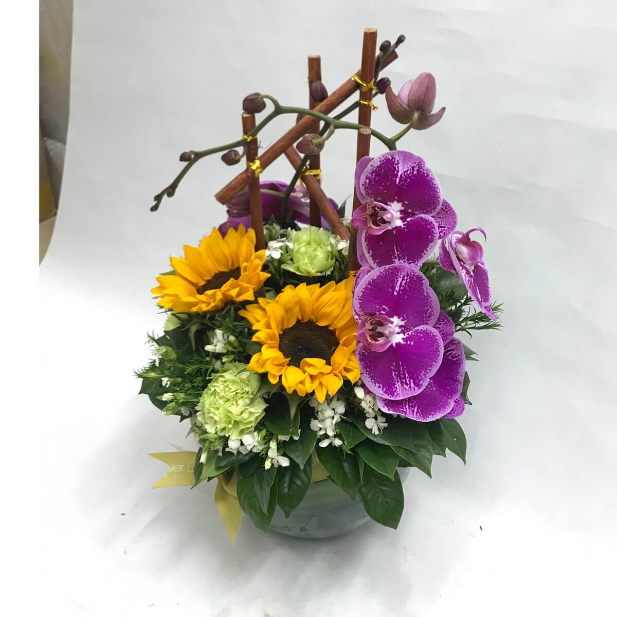 A cheerful mix of sunflowers, purple orchids, and green carnations in a round vase 向日葵與紫色蝴蝶蘭搭配綠色康乃馨,展現活潑亮麗風格,擺放於圓形玻璃花瓶中。