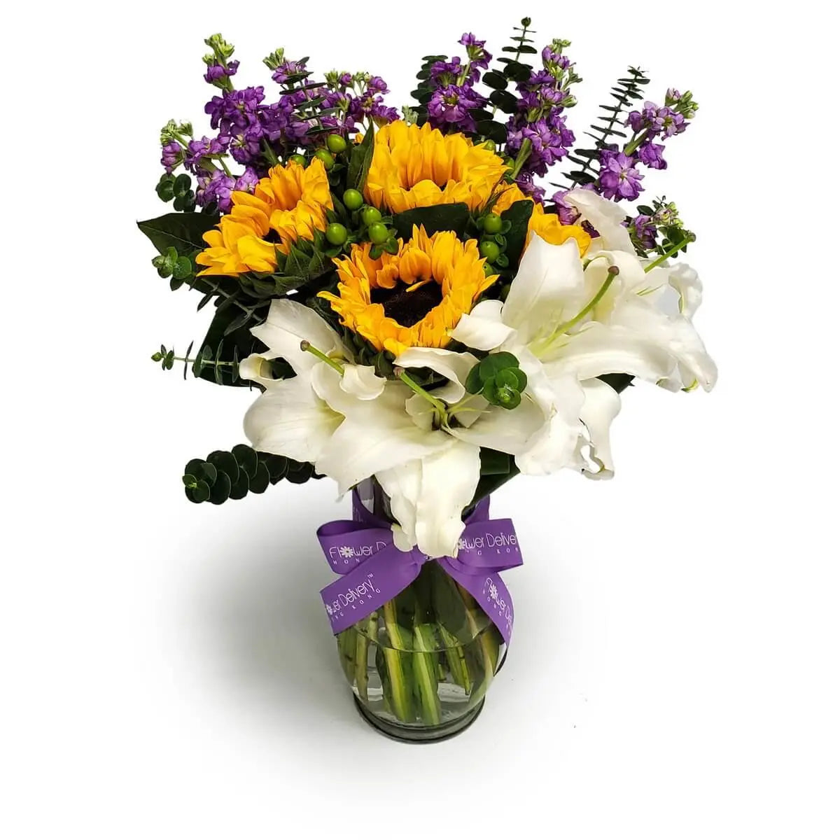 Arrangement of yellow sunflowers, white lilies, and purple stock with eucalyptus foliage in a clear glass vase tied with a purple ribbon | 向日葵、白百合與紫羅蘭配尤加利葉,透明玻璃瓶插並綁紫色絲帶。