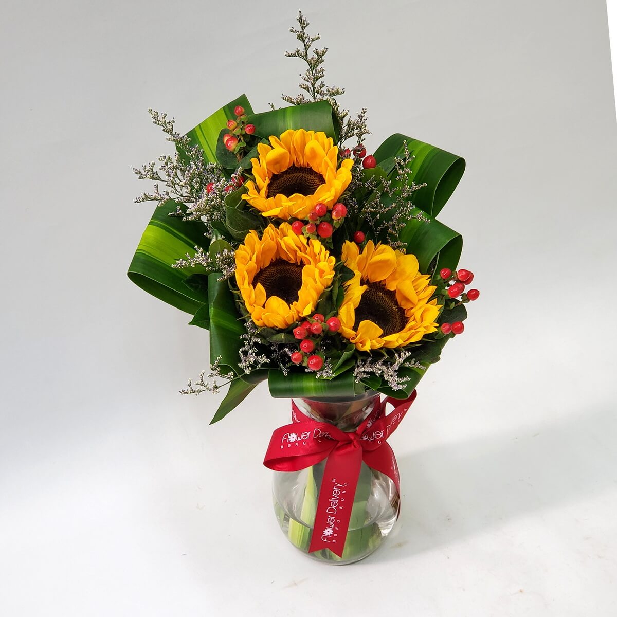 Vibrant sunflower trio with red hypericum berries and green foliage, presented in a clear vase – 三朵鮮豔向日葵、紅豆與綠葉構成的明亮花藝,透明玻璃花瓶裝飾,朝氣滿滿