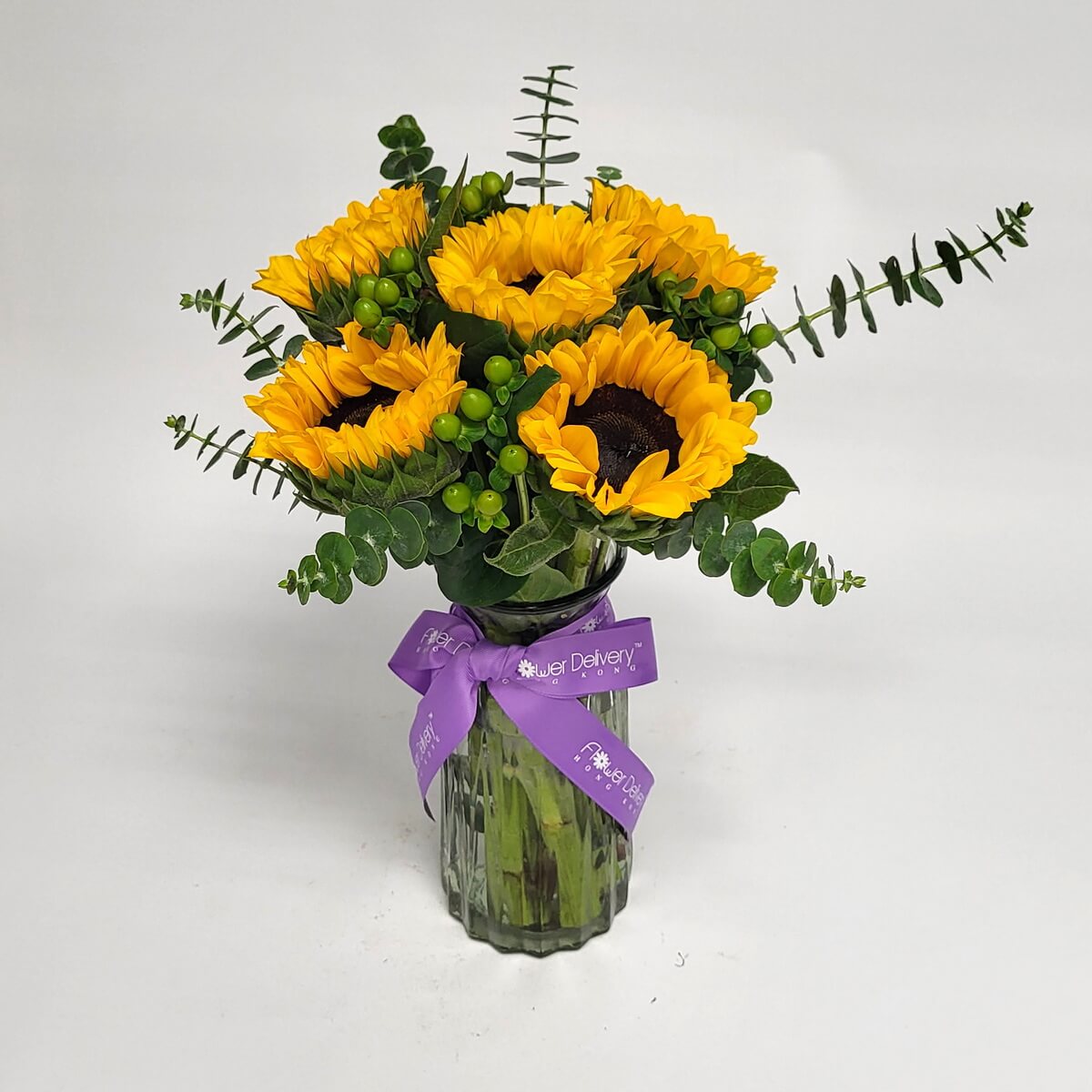 Cheerful sunflower bouquet with eucalyptus and berries, arranged in a clear vase with a purple ribbon 明亮向日葵與尤加利葉及漿果搭配,擺放在玻璃花瓶中,綁上紫色絲帶,洋溢朝氣。
