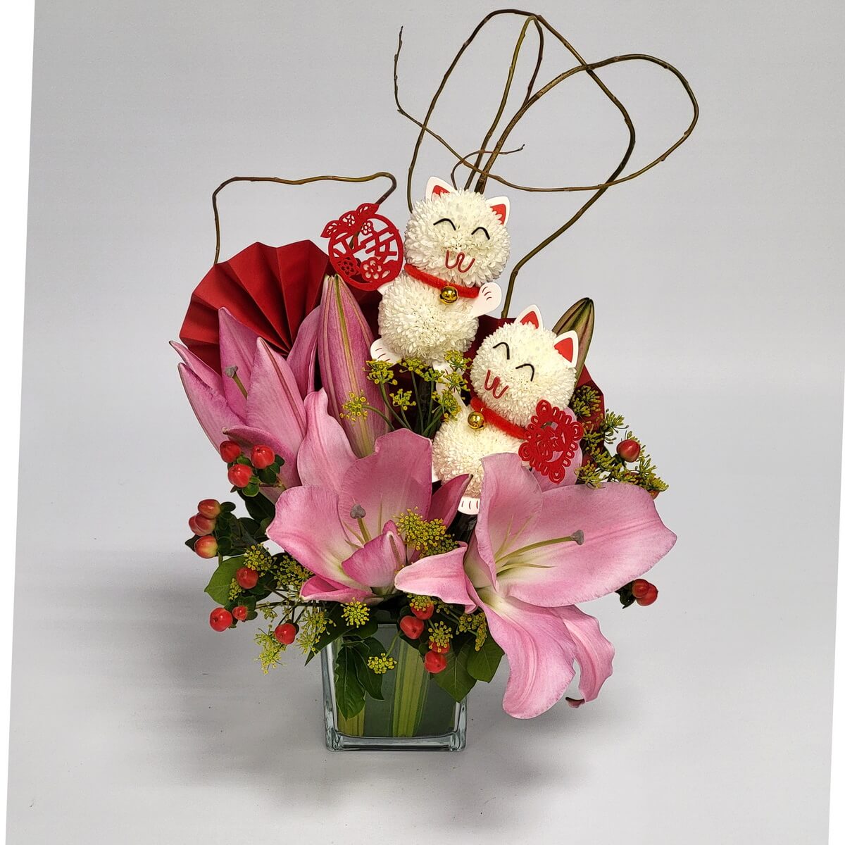 Arrangement of pink lilies, hypericum berries, yellow fillers, red paper fan, curly twigs, and festive cat decorations in glass vase 玻璃瓶插花設計,粉紅百合配紅色漿果、黃色配花、紅色紙扇、彎枝及兩個招財貓擺設,造型生動。