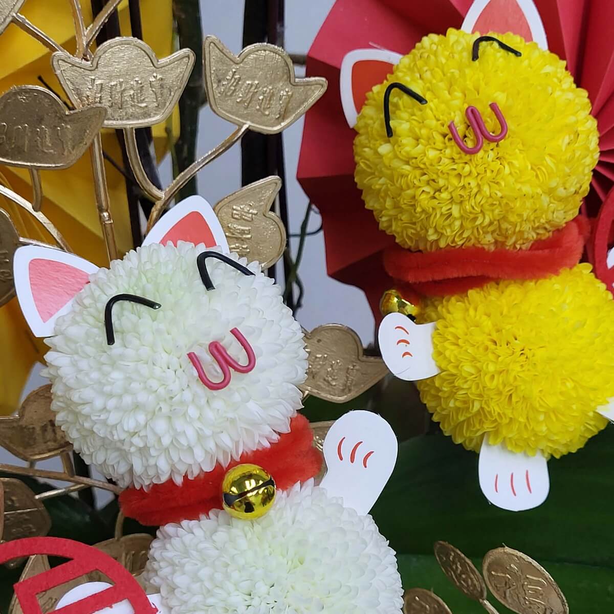 Detail view of white and yellow lucky cats made from button chrysanthemums, featuring red collars, gold bells, and paper ears amid gold coin decorations 白與黃乒乓菊招財貓造型特寫,配紅色頸圈、金鈴鐺及紙耳,襯以金幣裝飾背景
