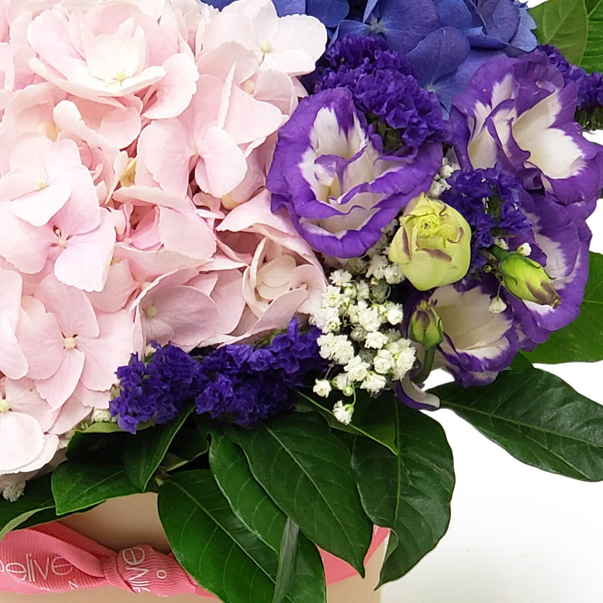 Detail view of a floral mix with soft pink hydrangeas, white and purple lisianthus, statice, and baby's breath, arranged with glossy green leaves - 淡粉繡球花配紫白洋桔梗、千日紫與滿天星,搭配亮綠葉的花藝細節圖