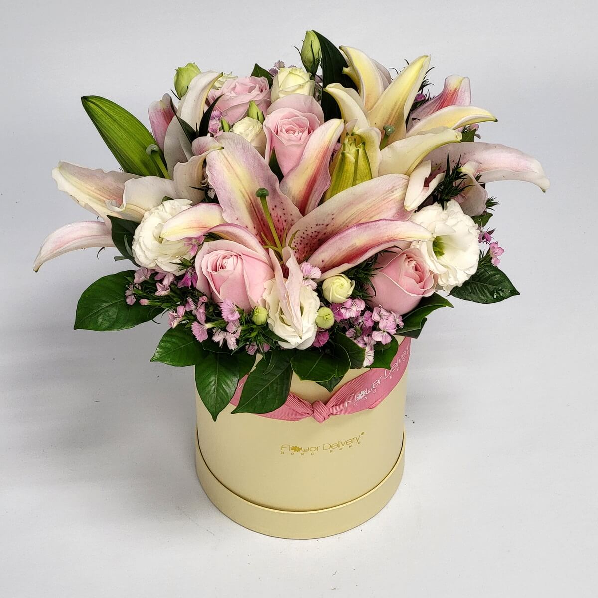 Arrangement of pink lilies, pale pink roses, white lisianthus, lavender filler flowers, and assorted greenery 粉紅百合搭配淡粉玫瑰、白色洋桔梗、紫色配花與多款綠葉,整齊設計於圓形花盒中。