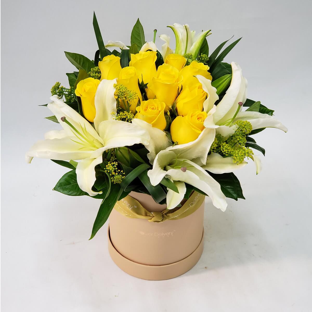 Arrangement of yellow roses and blooming white lilies in a beige round hat box with green fillers – 黃玫瑰與盛開白百合設計於米色圓形花盒中,搭配綠色配葉與花材,展現溫暖與純潔之美