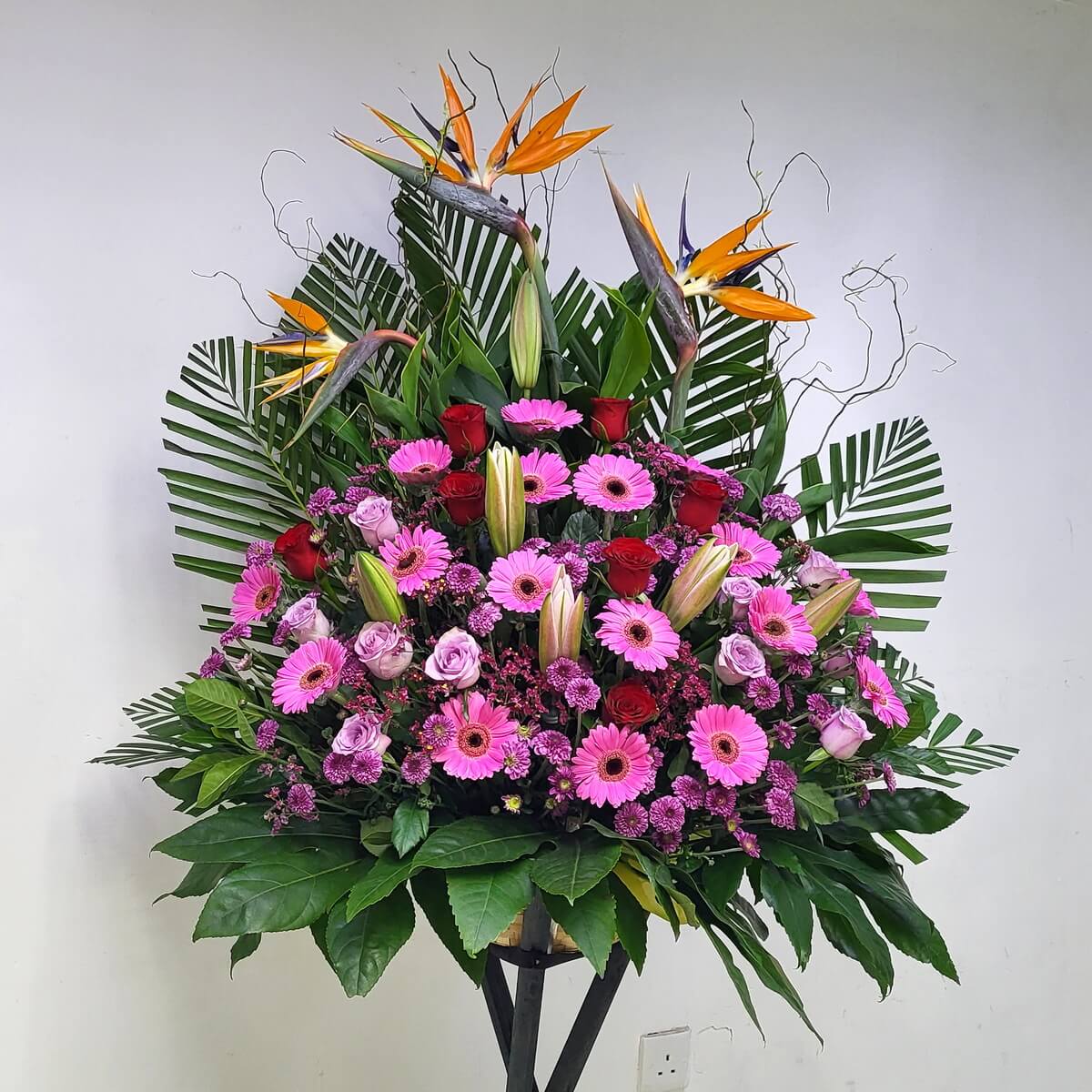 A tropical arrangement with bird-of-paradise, red roses, pink gerberas, lilies, and palm leaves.
熱帶風情花藝,融合天堂鳥、紅玫瑰、粉紅太陽花、百合與棕櫚葉,繽紛耀眼。